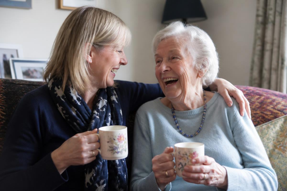 A woman and her mother laughing together over mugs of tea